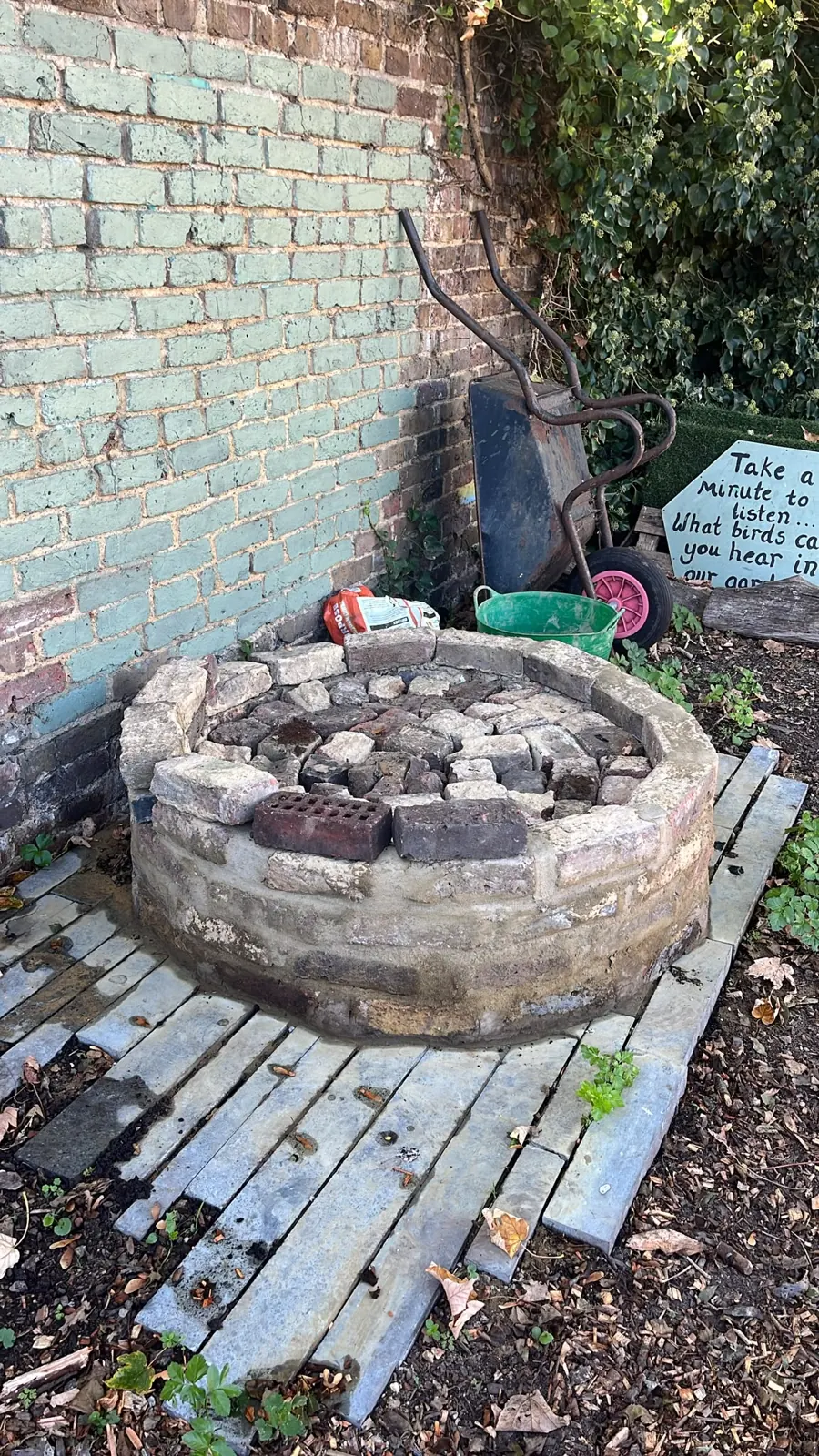 The round brick well on the decking, with a wheelbarrow and the listening board beside it